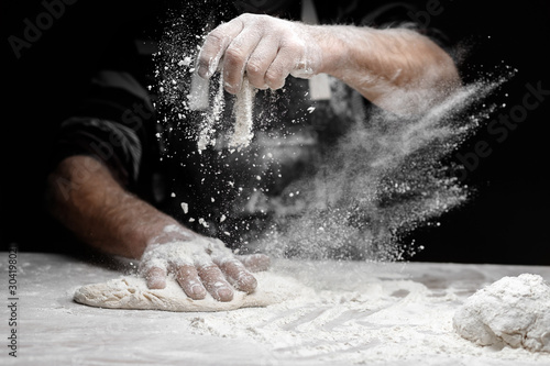 Fotografia White flour flies in air on black background, pastry chef claps hands and prepar