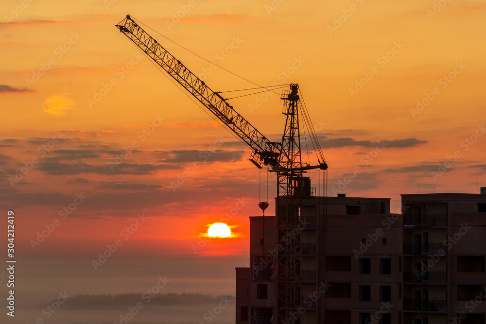 Tower crane on a construction site at sunrise with clouds. Unfinished high-rise building with luxury apartments in the city against the background of a forest in fog. Cityscape