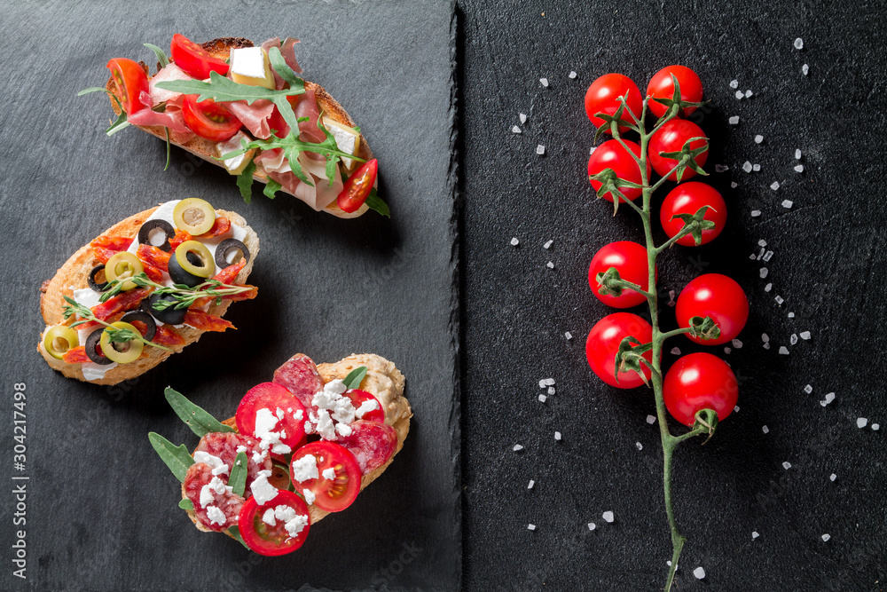 Traditional Bruschetta. Italian antipasti set with jamon, guanchial sausage, olives, cottage cheese, arugula and tomatoes on a black background Traditional Bruschetta. Italian antipasti set with jamon, guanchial sausage, olives, cottage cheese, arugula and tomatoes on a black background