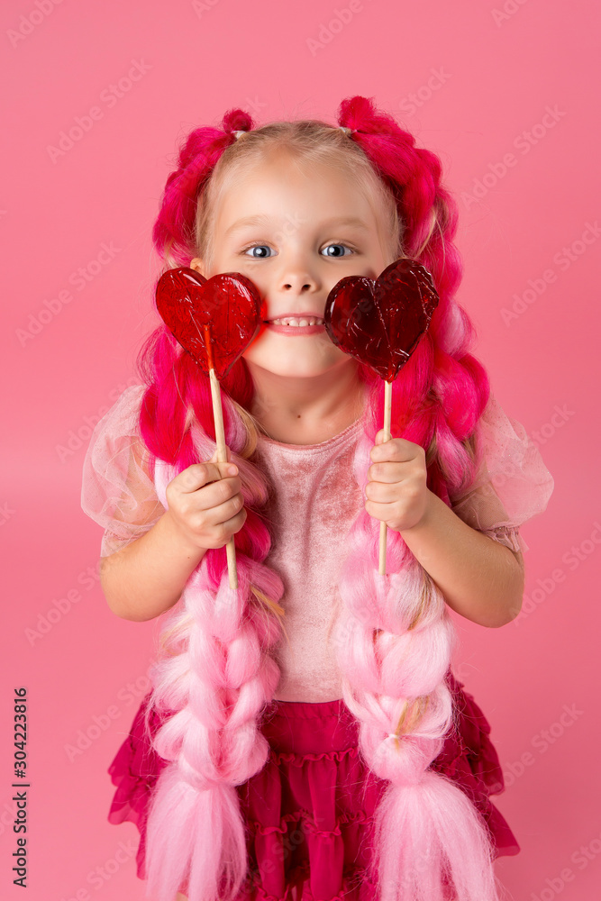 Fototapeta premium little girl with braids of pink kanekalon holds a heart-shaped Lollipop on a pink background. refusal of sweets. the concept of Valentine's day