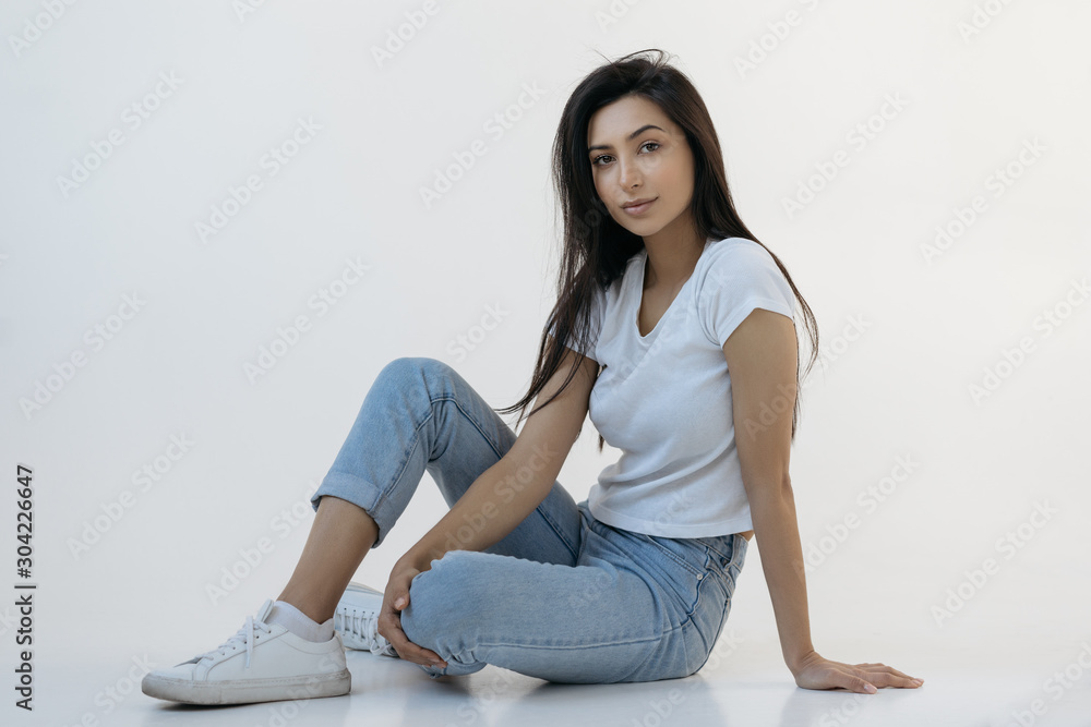 Portrait of young attractive woman with beautiful face wearing white t shirt and blue jeans sitting on the floor. Indian fashion model posing for pictures in studio isolated on white background Portrait of young attractive woman with beautiful face wearing white t shirt and blue jeans sitting on the floor. Indian fashion model posing for pictures in studio isolated on white background