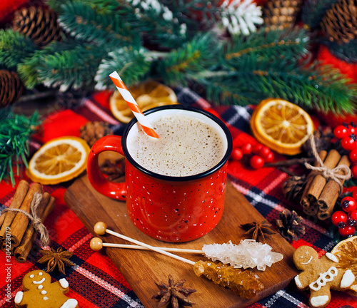 Christmas greeting card. Cacao drink in enamel cup with fir branches, dried orange slices, red berries, spices and gingerbread man on red fabric background.