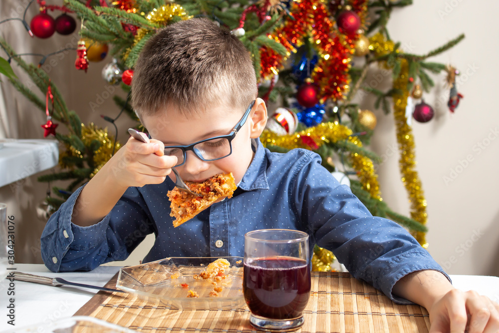 Chłopiec w okularach siedzi przy stole i ma buzię pełną jedzenia. Na talerzu leży lasagne a w tle pięknie ubrana choinka. Święta Bożego Narodzenia jedzenie. - obrazy, fototapety, plakaty 