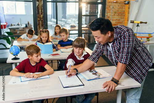 Pupils sit at desks and listens to the teacher while he explaining the topic of new lesson.