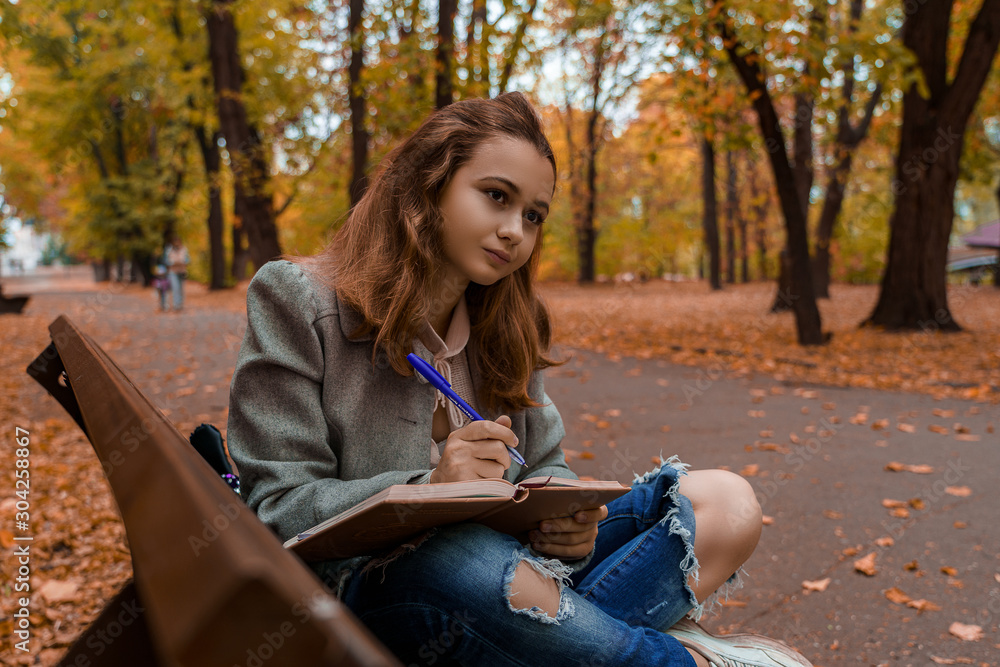 Obraz premium Beautiful teenage girl is sitting and writing in her notebook on the bench in fall park with yellow leaves