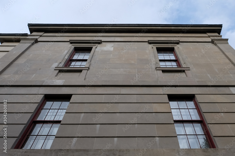 The exterior wall of the historic courthouse is made of blocks.The wall has four narrow tall windows.The ground upward view exposes blue sky with clouds. A formal style building with texture and lines The exterior wall of the historic courthouse is made of blocks.The wall has four narrow tall windows.The ground upward view exposes blue sky with clouds. A formal style building with texture and lines