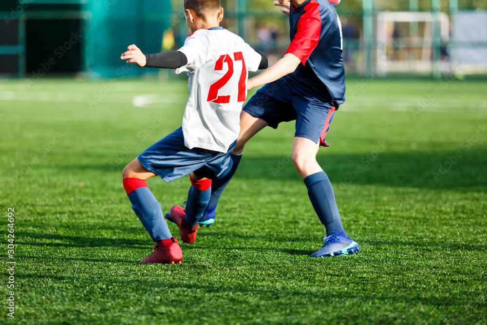 Obraz premium Boys in white and blue sportswear plays football on field, dribbles ball. Young soccer players with ball on green grass. Training, football, active lifestyle for kids concept 