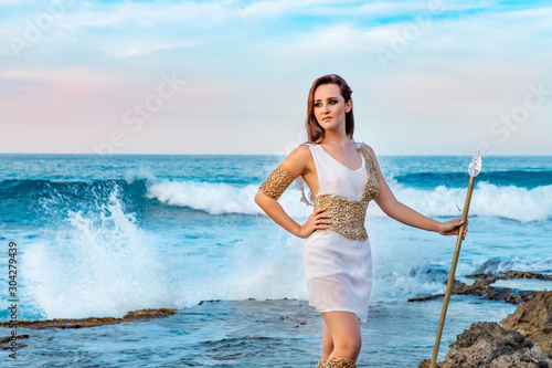 Fototapeta Naklejka Na Ścianę i Meble -  A young woman with long hair in a white and golden goddess outfit looking away hand on hip, with a spear on the ocean background, copy space