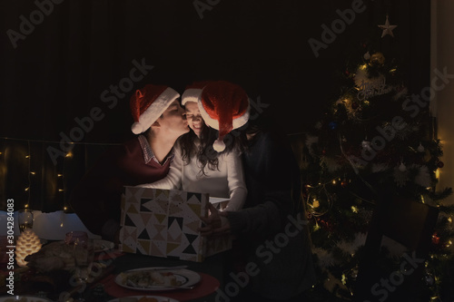 father and mother kiss daughter during open a gift present box together at christmas day night in living room that decorated with christmas tree for christmas festival day