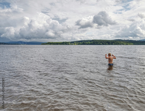 Young boy in blue swimming shorts stay in cold water of lake