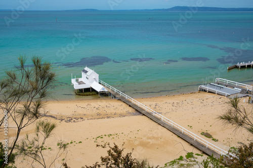 Long wooden pier extending over the beach into shallow turquoise bay water with white wooden shed at the tip