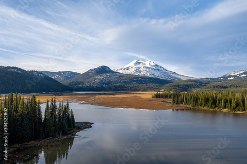Aerial view over tranquil lake surrounded by pine trees with picturesque snowy mountains in distance.