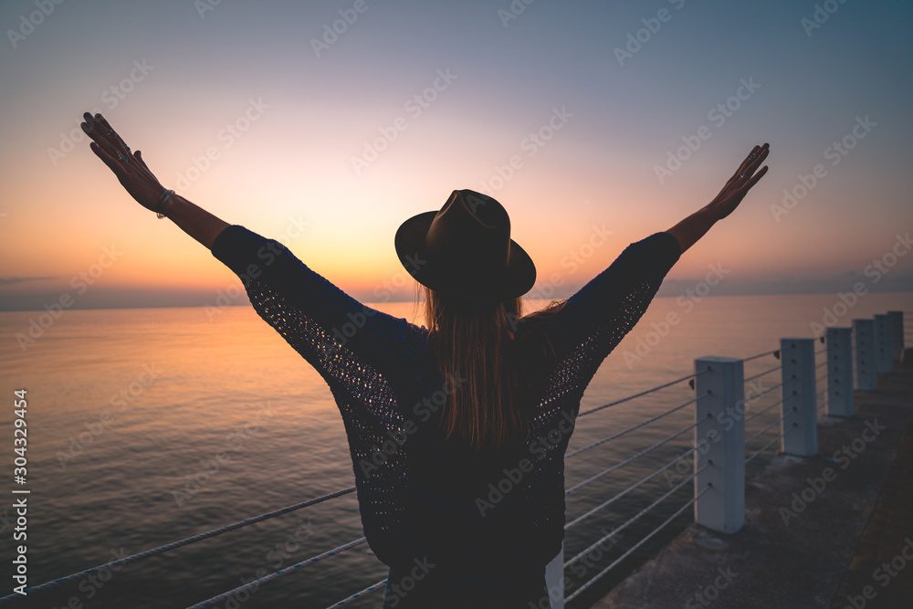Fototapeta premium One lonely carefree enjoy traveler woman in hat with open outstretched arms standing on sea pier at sunset. Happy freedom concept and enjoying life. Free people