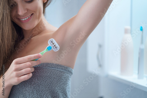 фотографія Happy attractive caucasian woman in towel removes hair under armpits with a female shaving razor in bathroom after shower