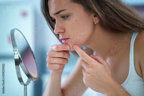 Upset stressed sad acne woman with problem skin squeezes pimple at home in front of a small round mirror photo