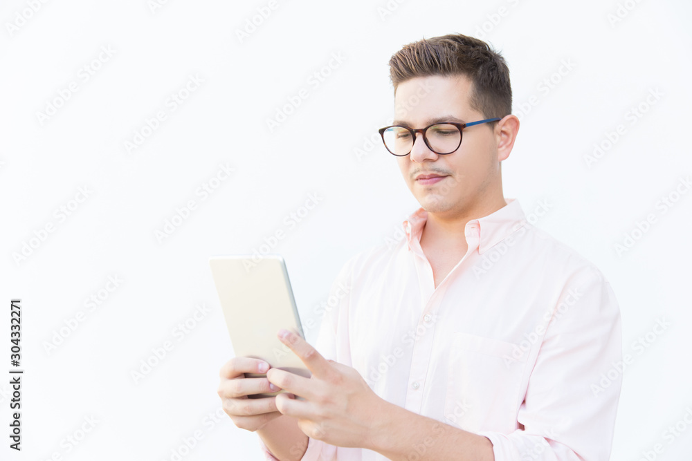 Focused guy in eyeglasses reading on tablet screen. Young man in glasses standing isolated over white background. Digital communication concept Focused guy in eyeglasses reading on tablet screen. Young man in glasses standing isolated over white background. Digital communication concept