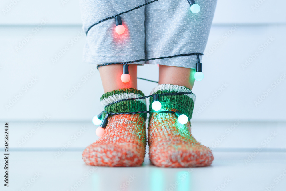 Female legs in warm knitted soft cozy christmas socks and garland with illuminated lights in wintertime at home. - obrazy, fototapety, plakaty 