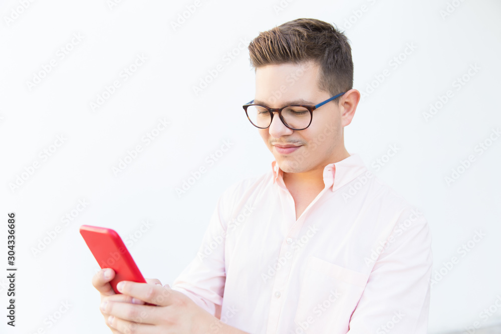 Focused guy in eyeglasses using smartphone, looking at screen. Young man in glasses standing isolated over white background. Texting message concept