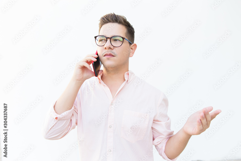 Sad disappointed guy in eyeglasses talking on cell and looking away. Young man in glasses standing isolated over white background. Digital communication concept Sad disappointed guy in eyeglasses talking on cell and looking away. Young man in glasses standing isolated over white background. Digital communication concept