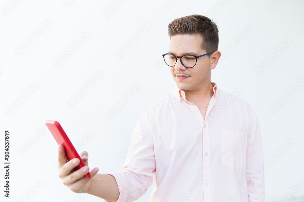 Serious focused guy in eyeglasses reading message on mobile phone. Young man in glasses standing isolated over white background. Wireless communication concept