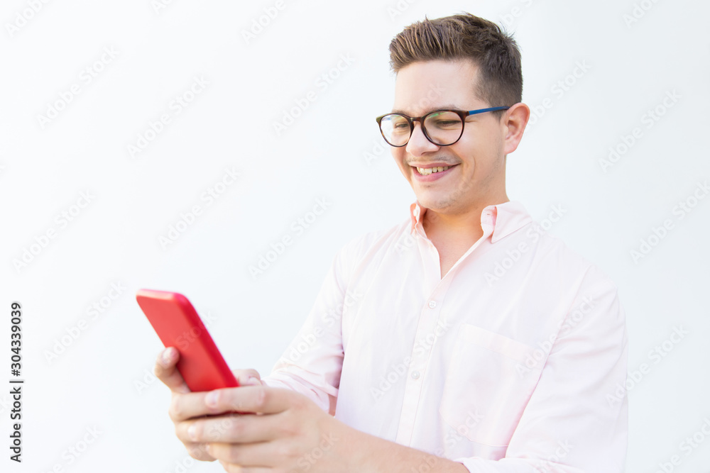 Positive joyful guy in eyeglasses reading message on phone and laughing. Young man in glasses standing isolated over white background. Good news concept