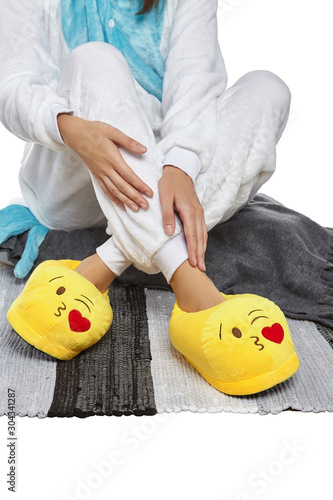 Cropped medium shot of a girl in white velour pyjamas and yellow plush house slippers made as kissing heart emoji. The girl is sitting on the striped carpet and a gray plaid.