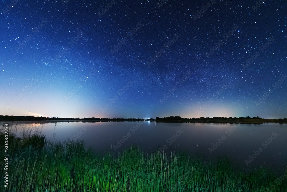 A magical starry night on the river bank with a large tree and a milky way in the sky and falling stars in the summer.