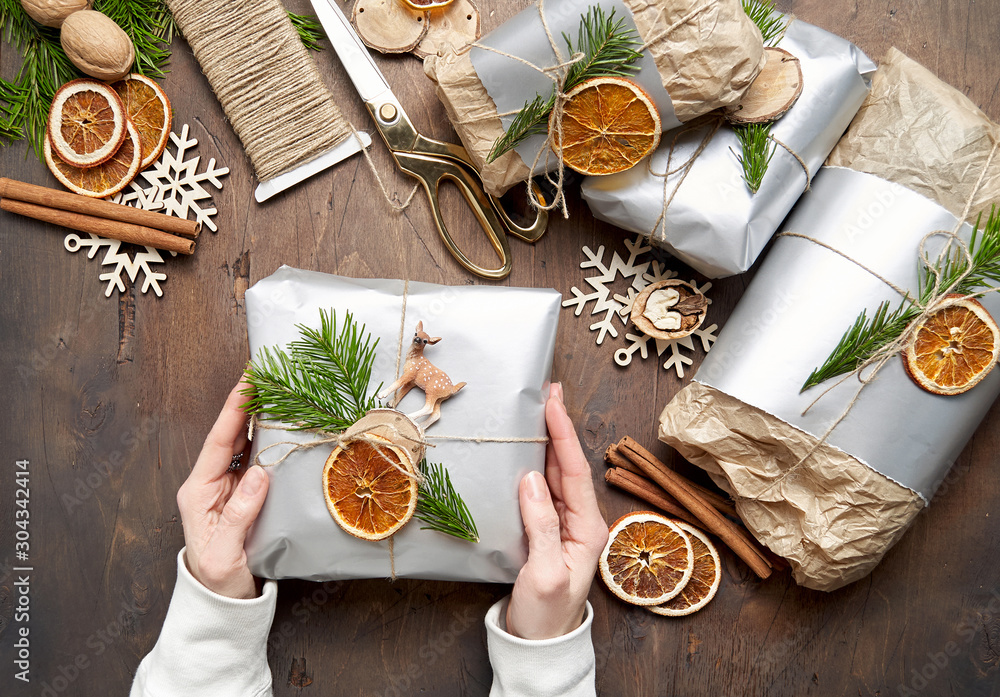 Woman is packaging christmas present handmade gift boxes decorated with silver paper with dried orange slices