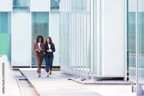Full length portrait of smiling businesswomen walking with tablet. Cheerful young women wearing formal wear looking at tablet during stroll. Business concept