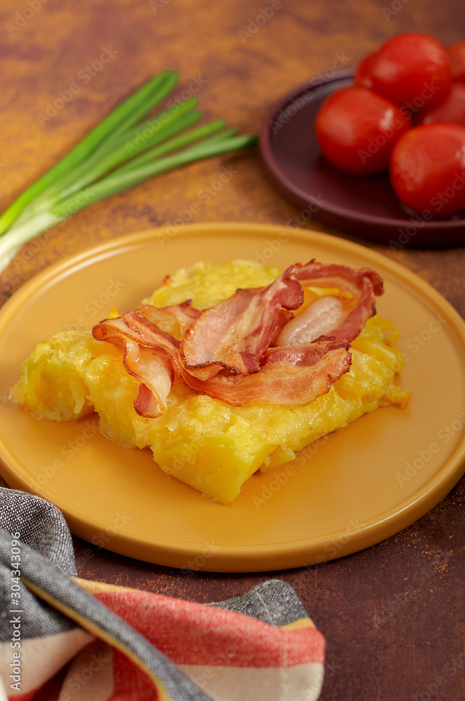 Cheesy potato bake with fried bacon, pickled tomatoes and green onion on yellow plate