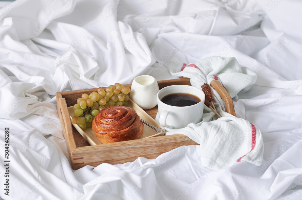 Breakfast on white bed sheets in hotel room. Close up of wooden tray with coffee, bun, jam and grape