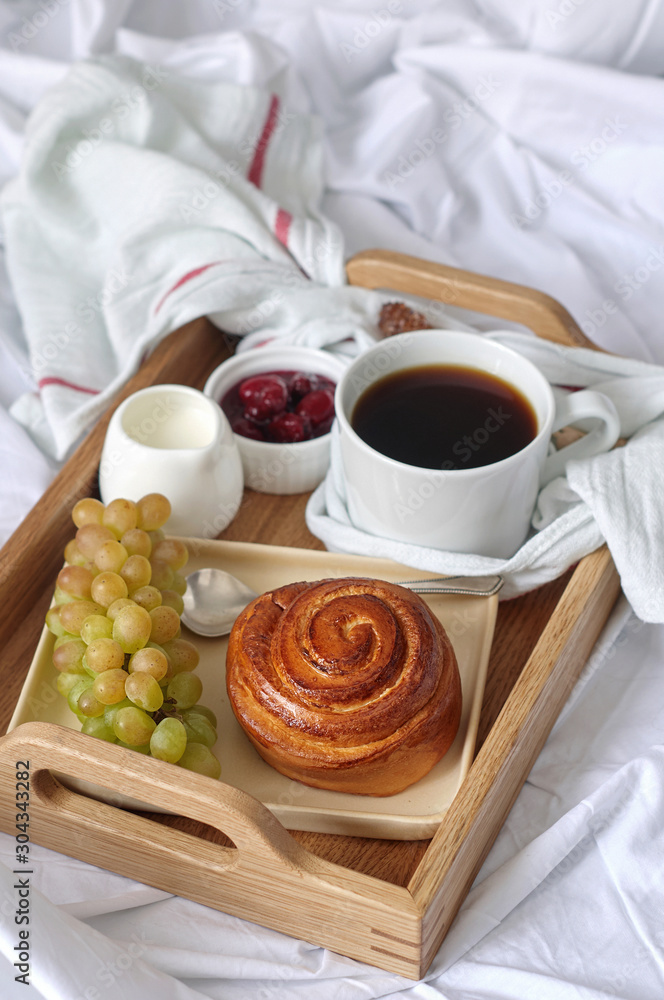 Breakfast on white bed sheets in hotel room. Close up of wooden tray with coffee, bun, jam and grape