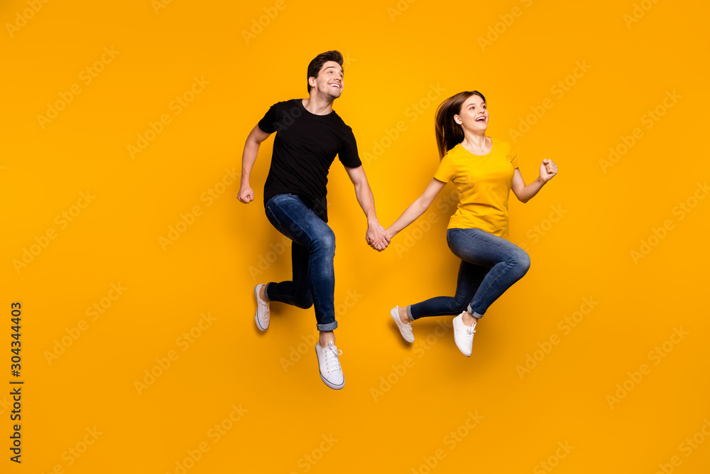 Full size photo of sporty guy and lady couple jumping high holding hands pair speed marathon participants wear casual jeans t-shirts isolated yellow color background