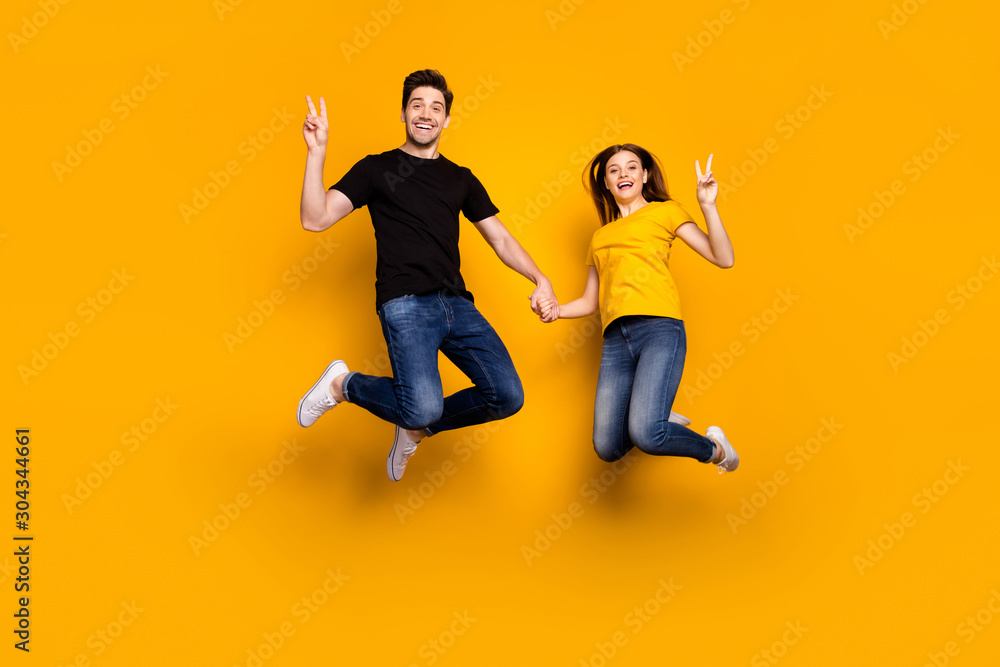 Full body photo of crazy guy lady couple jumping high holding arms showing v-sign symbols saying hi wear casual jeans black t-shirts isolated yellow color background