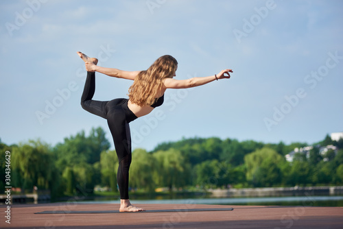 Sporty woman in black sportswear doing yoga at city lake. Green park on the background. Natarajasana exercise, Lord of the Dance pose, beautiful long hair, side view, full length