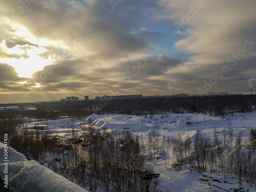 A winter landscape with tall snowdrifts, bare trees, low gray clouds in a blue sky and the sun shining through them and tall houses on the horizon