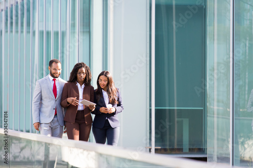 Three smiling coworkers looking at tablet during stroll. Cheerful business partners walking on street during sunny day. Business concept