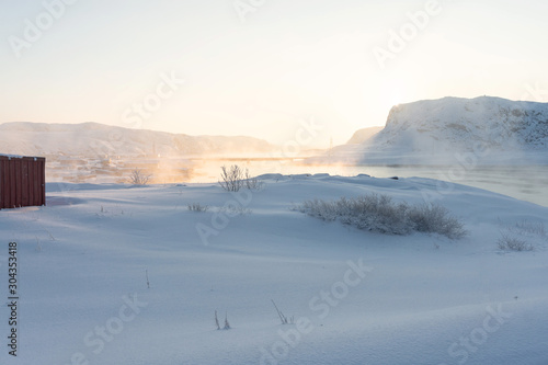 snow-clad shore of the bay lit by the rays of the orange sun at dawn beyond the Arctic Circle. dense fog rises from the water on a frosty winter day © Александр Коновалов