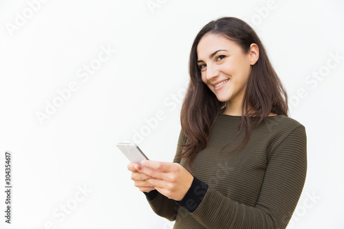 Smiling satisfied cellphone user texting message. Young woman in casual with mobile phone standing isolated over white background. Communication concept