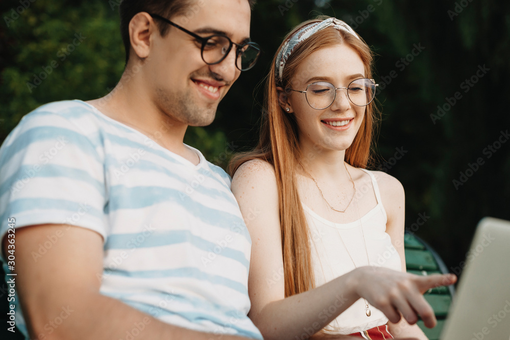 Charming young couple looking at their laptop smiling while freelancing outdoor in the park on a beach . Young programsit couple working outside. Charming young couple looking at their laptop smiling while freelancing outdoor in the park on a beach . Young programsit couple working outside.