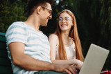 Beautiful young caucasian couple looking to each other laughing while sitting on a beach with a laptop on the legs in the park.