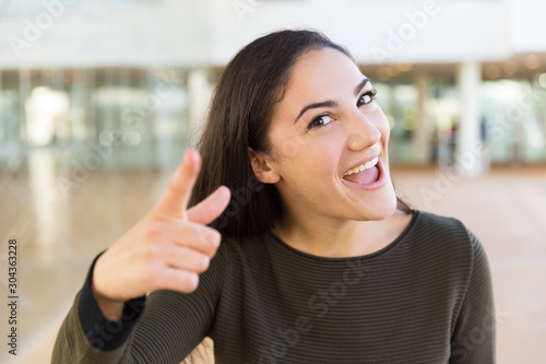 Excited joyful beautiful woman gesturing with hand and index finger, smiling and shouting. Young woman in casual posing indoors with glass wall interior in background. New idea concept