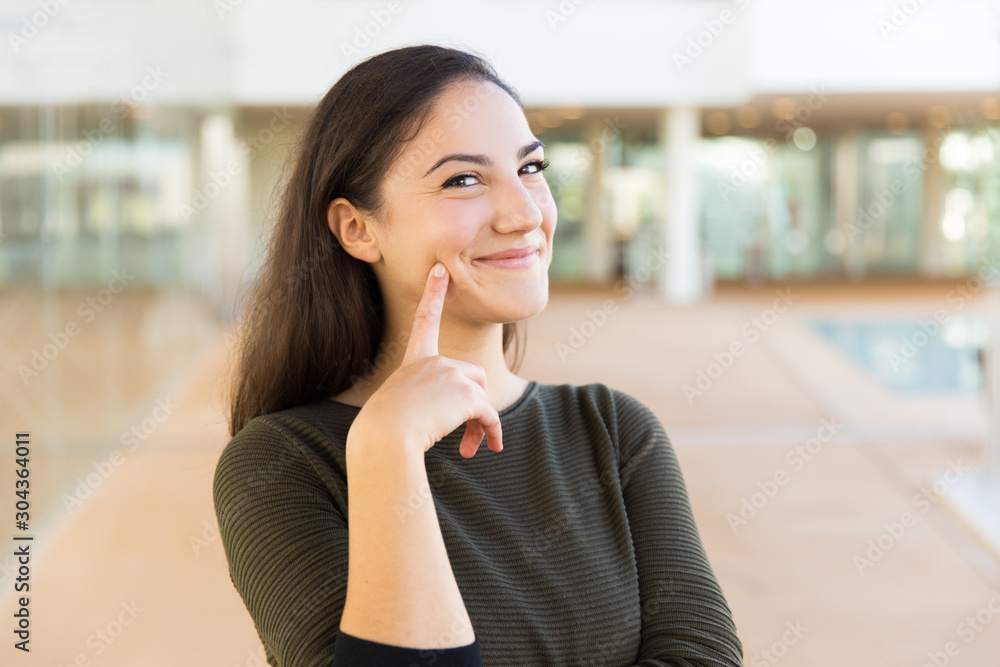 Happy cheerful beautiful woman touching face with index finger. Young woman in casual posing indoors with glass wall interior in background. Skin care or female portrait concept Happy cheerful beautiful woman touching face with index finger. Young woman in casual posing indoors with glass wall interior in background. Skin care or female portrait concept