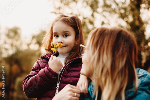 Lovely little girl looking at camera smiling while smiling a little yellow flower while her mother is looking at her outdoor.