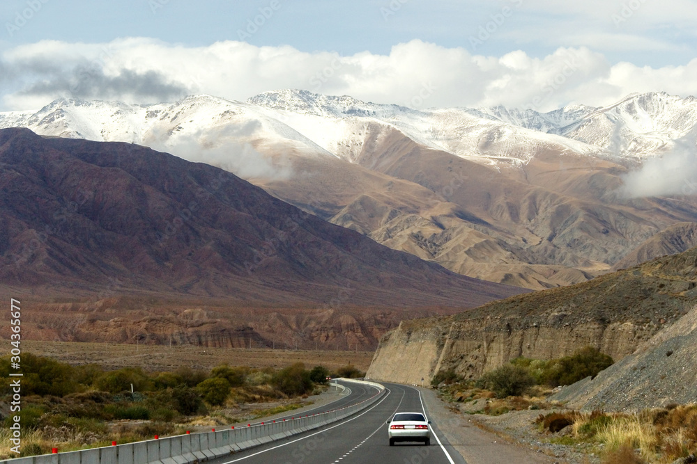 Highway with a car on a background of high mountains, some of which are covered with snow, Kyrgyzstan