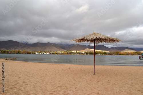 A lonely beach umbrella made of reeds on a empty beach. In the background there are gray clouds over the village and the mountains. Bad weather on vacation. Closing of the beach season  Kyrgyzstan