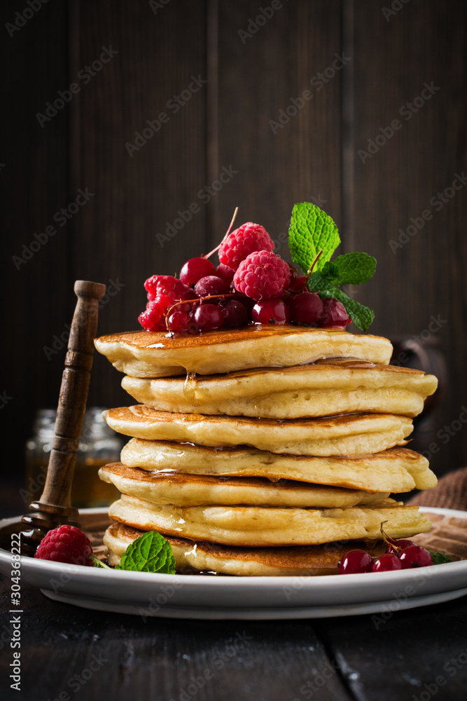 Stack of homemade little pancakes with honey, fresh raspberries and red currants on an old wooden background. Selective focus. Stack of homemade little pancakes with honey, fresh raspberries and red currants on an old wooden background. Selective focus.
