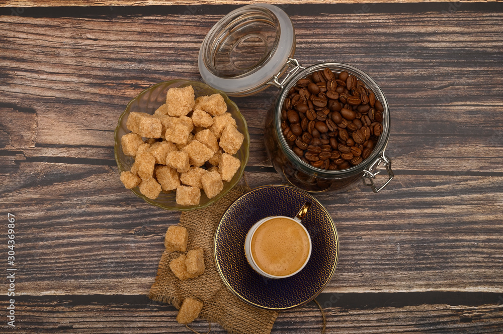 A Cup of coffee, coffee beans in a glass jar and pieces of brown sugar on a wooden background.