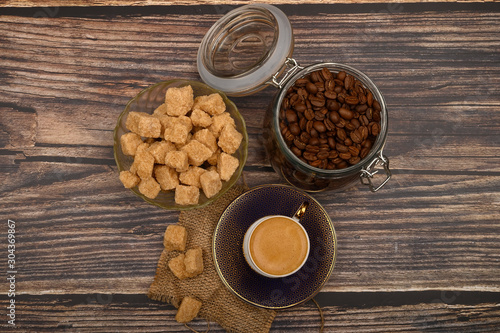 A Cup of coffee, coffee beans in a glass jar and pieces of brown sugar on a wooden background.