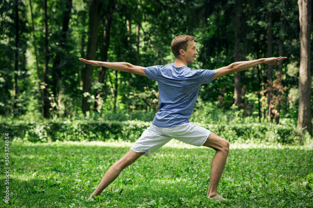 Young attractive man in a gray T-shirt and sweatpants doing yoga warrior pose in the park. He spread his legs wide, arms outstretched to the sides, looking to the side.